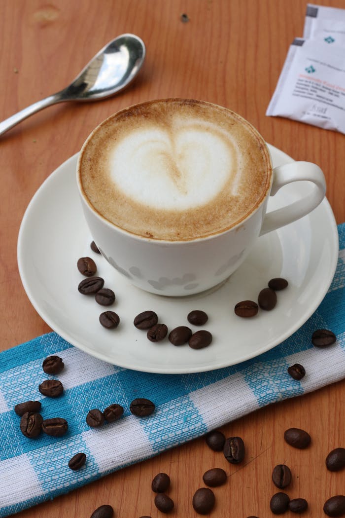 A coffee cup with heart-shaped latte art surrounded by coffee beans on a checkered cloth.